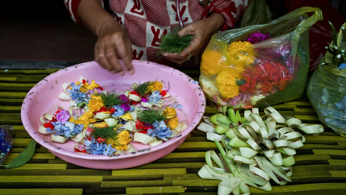 Hands assembling small palm-leaf offerings of flowers on a woven mat