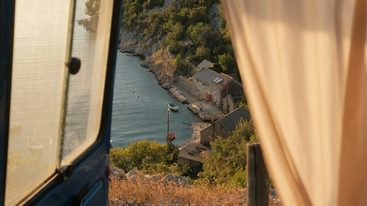 A cove with a small stone harbour seen through the half-open window and curtain of a camper van parked above the sea