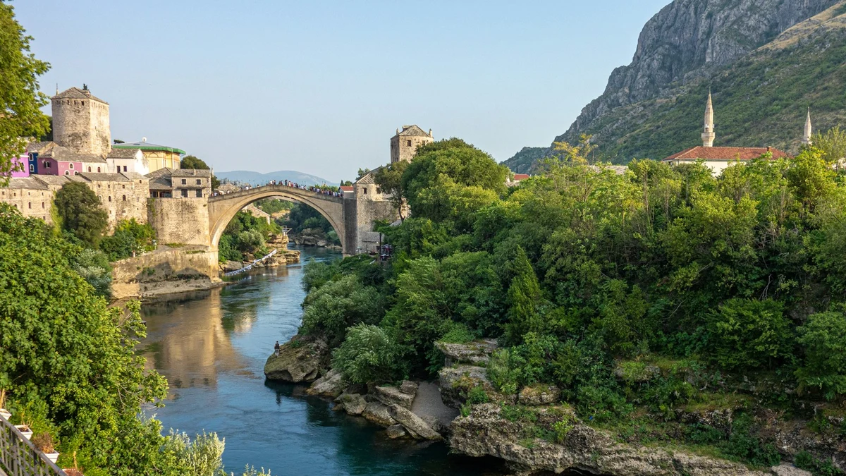 The Old Bridge of Mostar arching over the green Neretva river, with stone towers, a minaret and a steep mountain behind