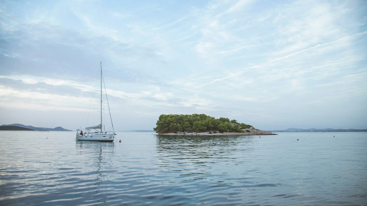 A white sailing yacht at anchor on flat morning water next to a small wooded island, with low coastline on the horizon