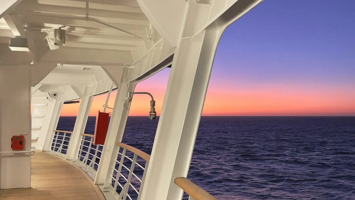 A covered promenade deck on a cruise ship at dusk, with white railings and a pink sunset over the open sea