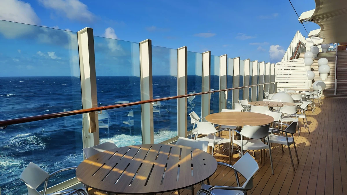Empty round tables and white chairs on a sunny open deck behind a glass windbreak, with deep blue sea beyond