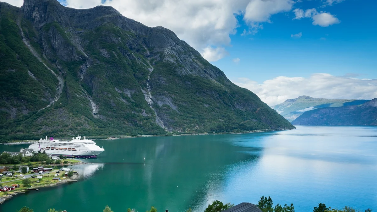 A white cruise ship moored at a small village pier at the foot of steep green mountains in a Norwegian fjord