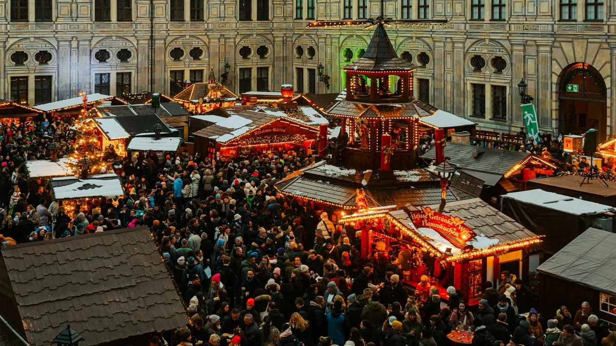 A crowded Christmas market seen from above, with wooden stalls, snow on the roofs and strings of warm lights