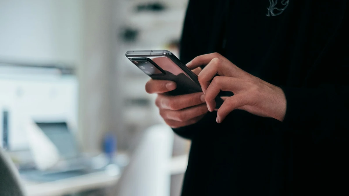 Two hands holding a smartphone and typing, with a blurred bright office in the background