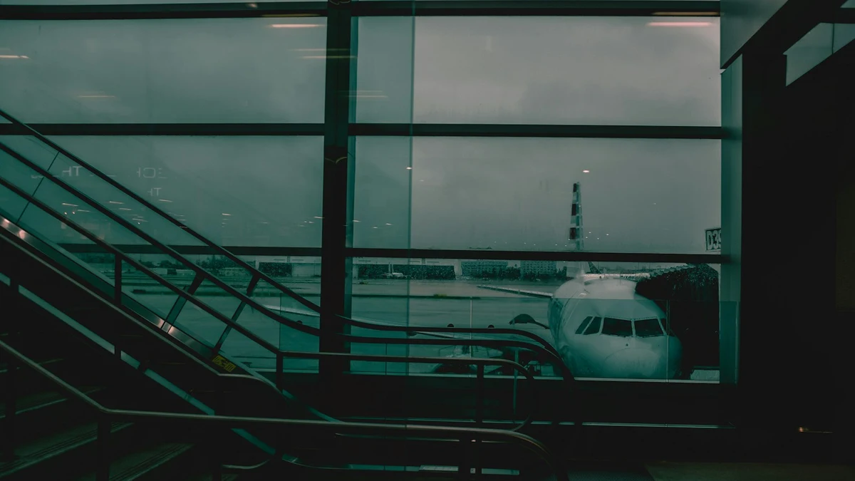 A dim airport terminal with an escalator and large windows, a parked passenger jet visible on the apron outside