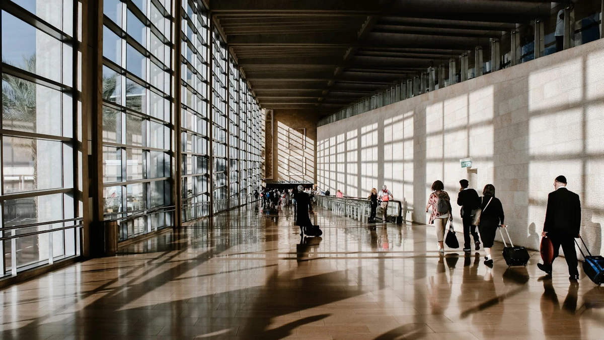 Travellers with suitcases crossing a sunlit airport terminal beside a wall of tall windows