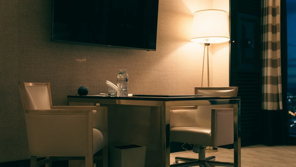 A desk and two chairs in a hotel room at night, lit by a floor lamp, city lights beyond the curtain