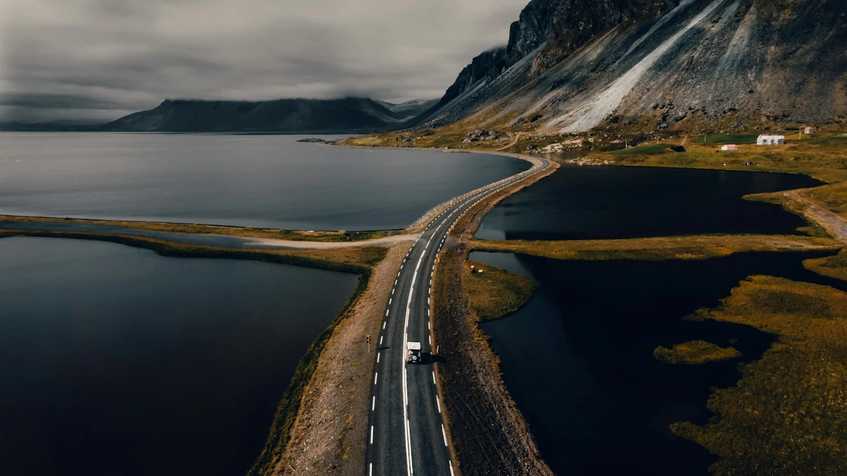A two-lane road on a causeway between dark lagoons, curving towards a steep mountain under a grey sky