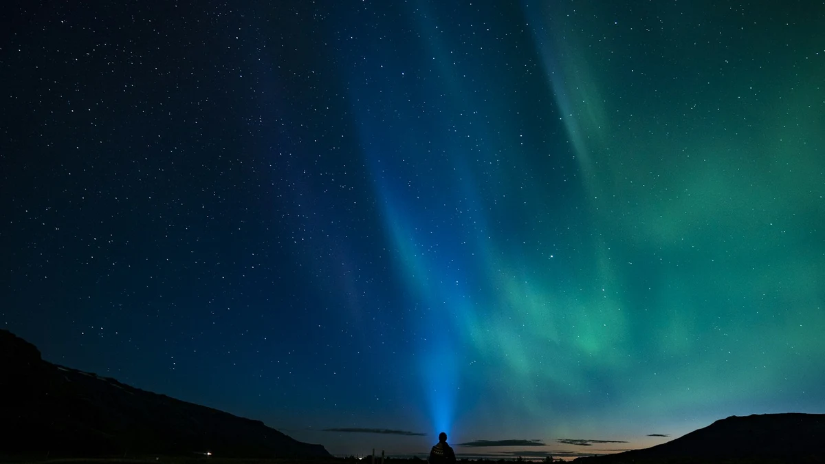 A lone figure with a head torch standing on a dark plain under blue and green northern lights