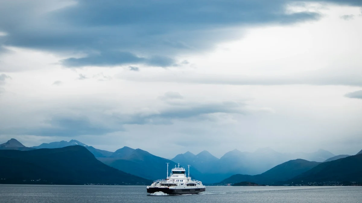 White car ferry crossing a wide fjord under heavy grey clouds, with blue mountain ridges behind