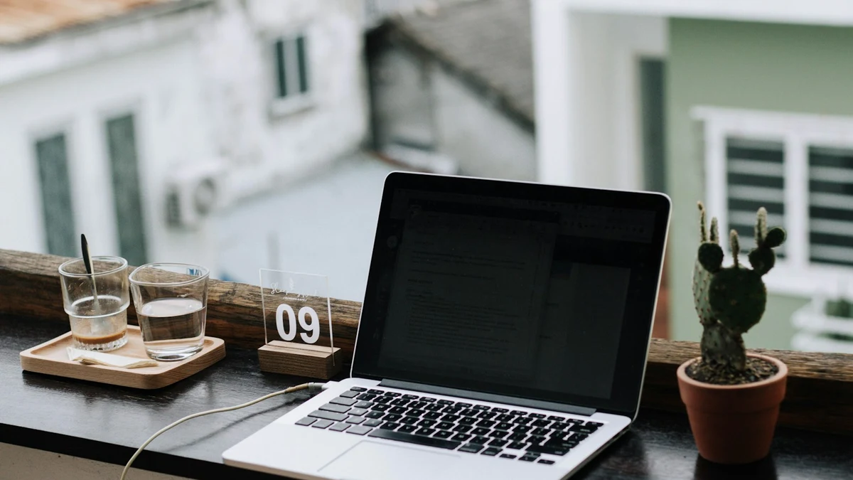 Laptop on a windowsill table with glasses and a small cactus, street view behind