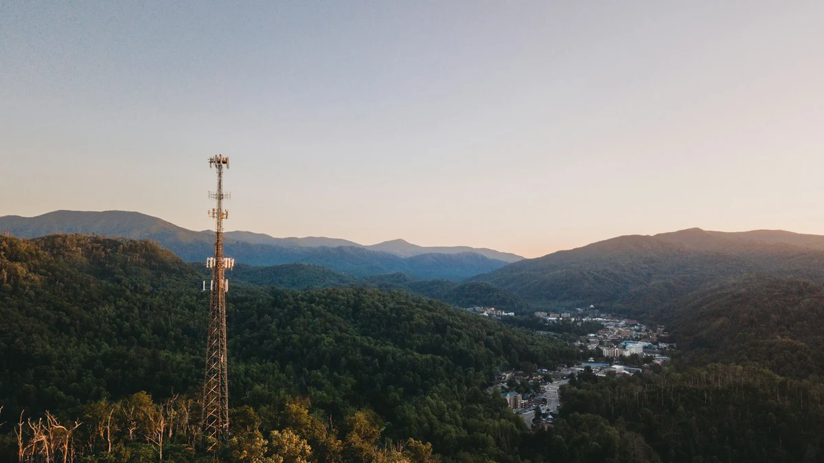 A cell tower on a forested ridge above a small town in a mountain valley at dusk