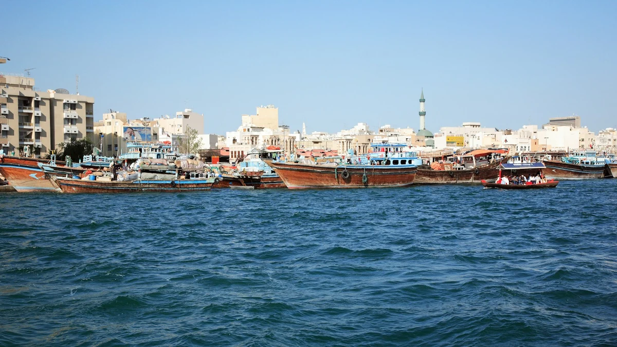 Wooden dhows moored along Dubai Creek in front of the low-rise buildings and a minaret of old Deira under a clear sky