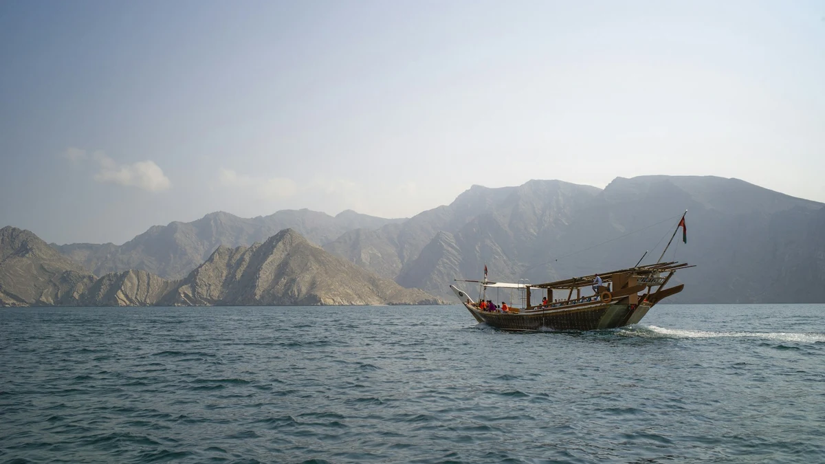 A traditional wooden dhow cruising across calm water in a Musandam fjord, with bare mountain ridges behind