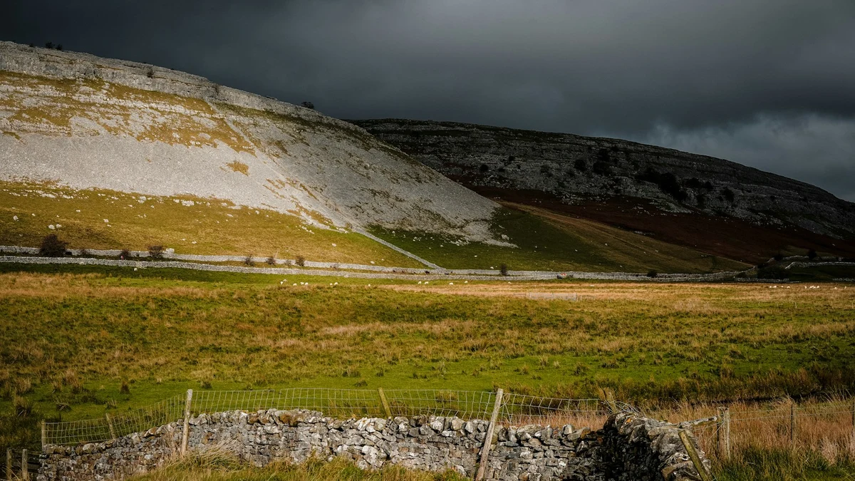 A limestone scar above rough pasture divided by dry stone walls under a dark storm sky